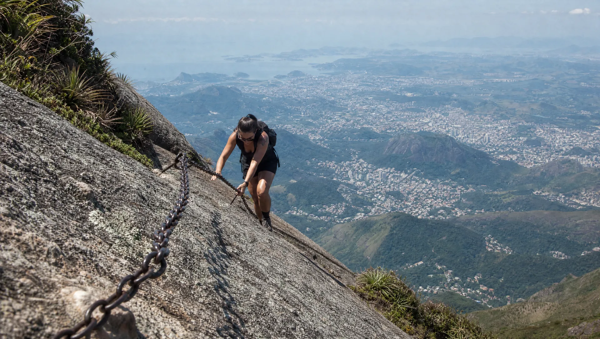 Trilheiro subindo lajedo íngreme com correntes no Escalavrado, Teresópolis ao fundo